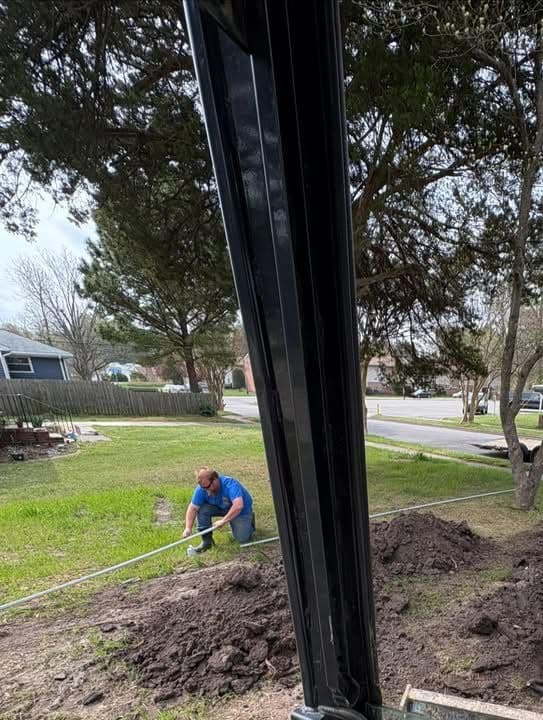 Man digging in yard, surrounded by trees and grass, with a residential area in the background.