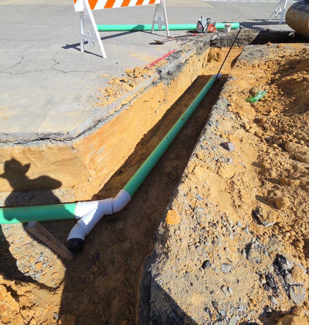 Excavated trench with green and white plumbing pipes in road construction site.
