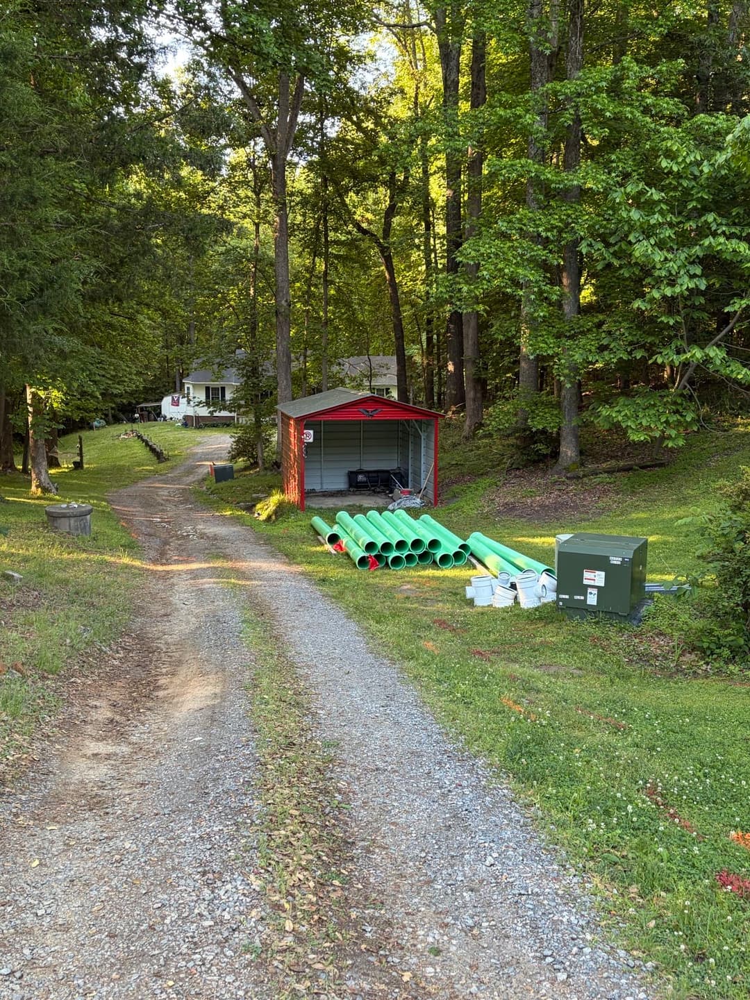 Gravel driveway leading to a storage shed and green PVC pipes in a wooded area.