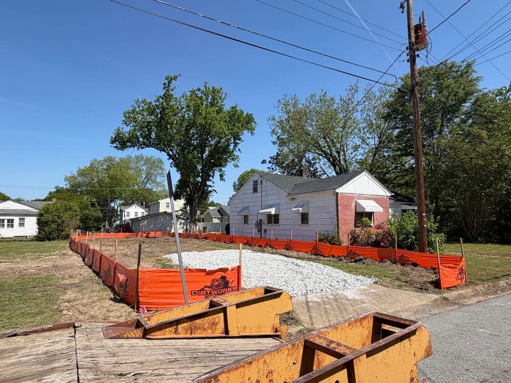 Residential construction site with gravel and orange fencing, featuring a nearby house.