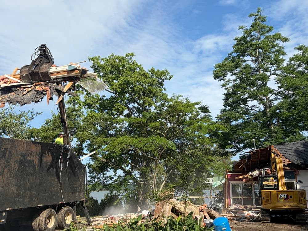 Construction workers operating heavy machinery during home demolition near a lakeside.